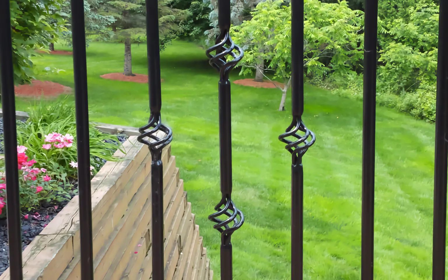 Black decorative basket twist aluminum balusters installed in a deck railing with lush green garden in the background