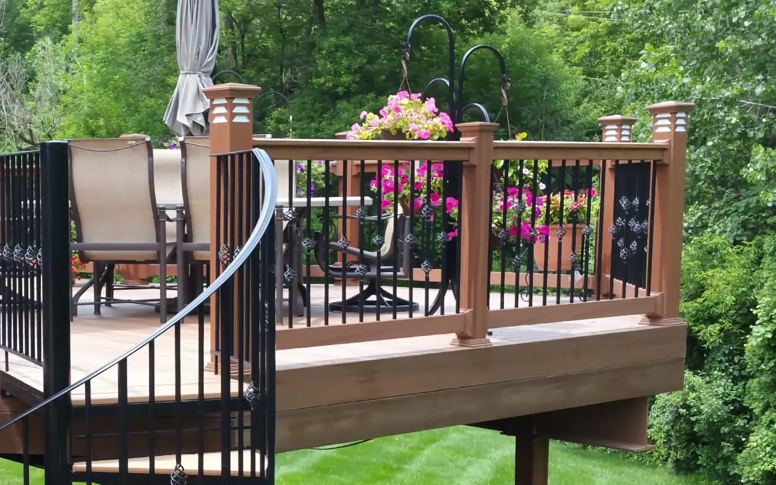Raised composite deck with black aluminum railing round balusters spiral staircase and copper post caps surrounded by flowers and greenery